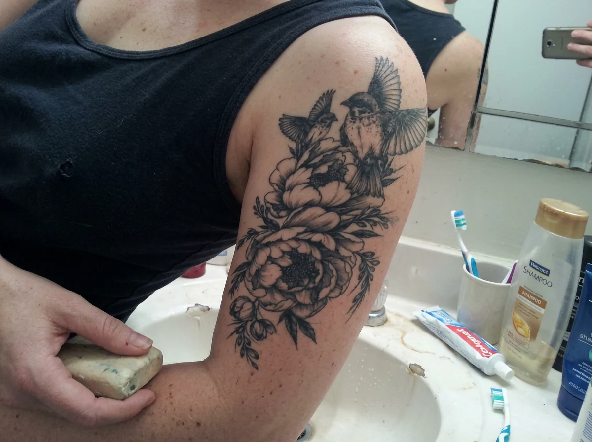Close phone photograph of a woman's Bird Blossom temporary tattoo on her upper arm beside a bathroom sink